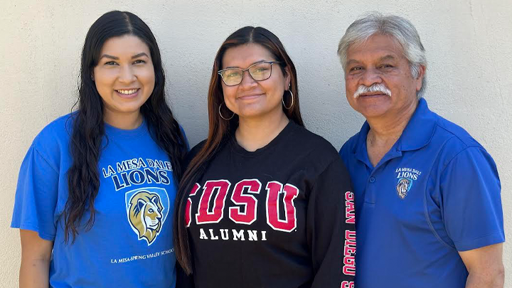 A young woman with glasses and a black SDSU Alumni sweatshirt stands between her sister and her mustachioed father, both wearing blue La Mesa Dale clothing.