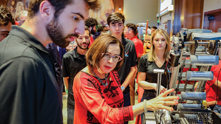 The SDSU president, in glasses and wearing a red and black dress, is examining a mechanical device as a bearded young man to her right looks on. In the background are three other young people, also watching her.