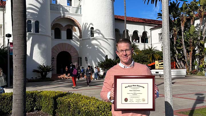 A middle-aged man wearing a salmon-colored pullover is holding a framed diploma with the words San Diego State University at the top. In the baqckground is a low bush, a Spanish Mission-style building and a half-dozen people, standing or talking.