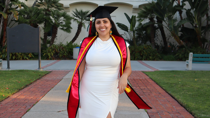 Daisy Lopez is photographed in a white dress and her SDSU cap and gown outside of the campus