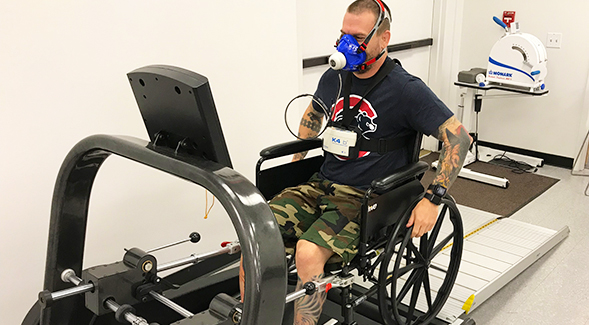 Exercise physiology graduate student Eric Bartholomae demonstrates the mobile metabolic cart which measures calories burned while pushing a wheelchair on the roller system treadmill.