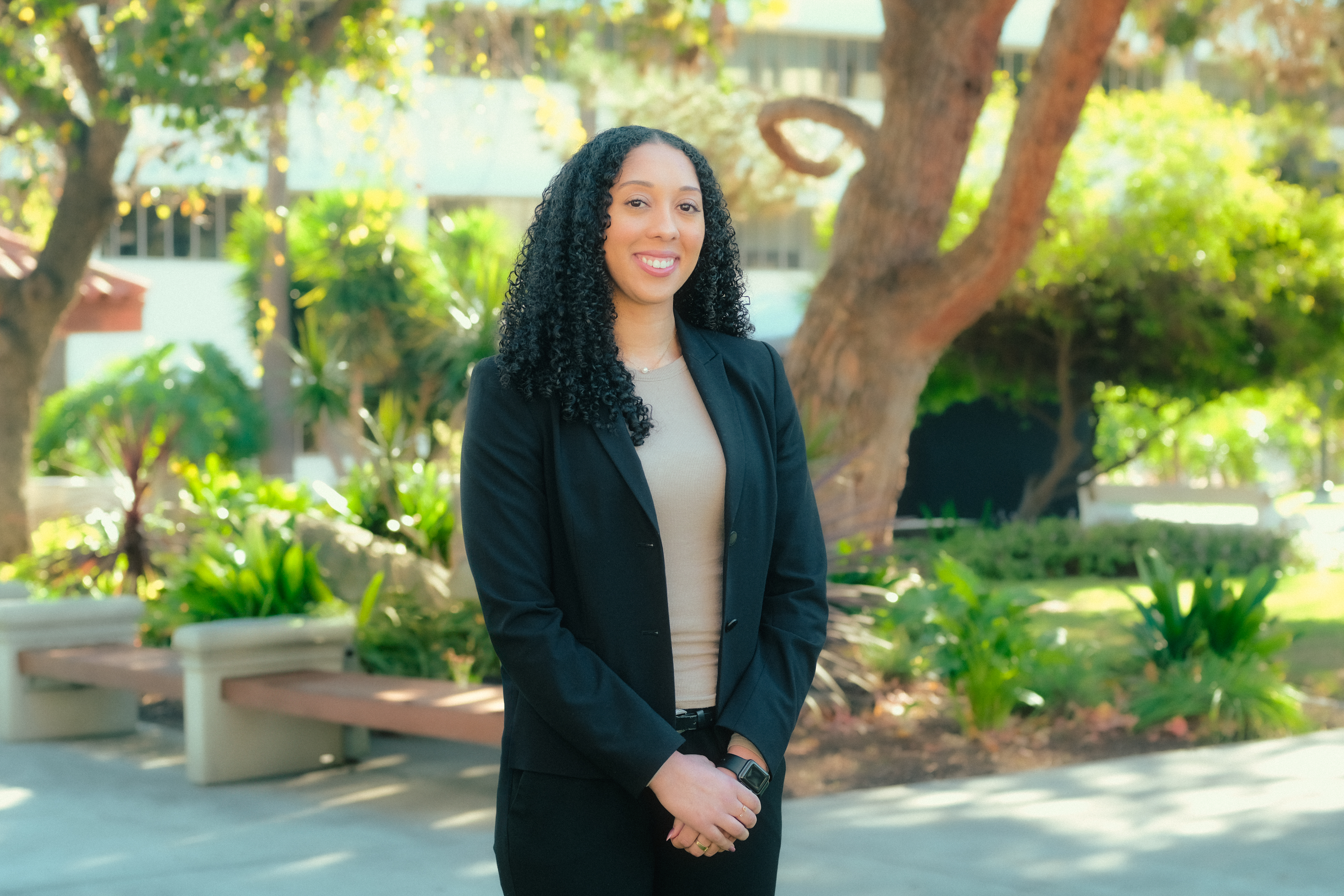Doctoral student Breanna Leone is smiling, her hair falling over her right shoulder and hands clasped in front of her. She is standing on a sidewalk outside SDSU's Lamden Hall. Behind her are a bench, a small garden and green space and a tree