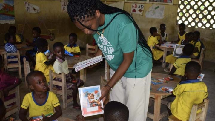 A young woman in a teal t-shirt with white slacks is standing in a classroom amid a cluster of tables and chairs. She is handing a publication to a child.