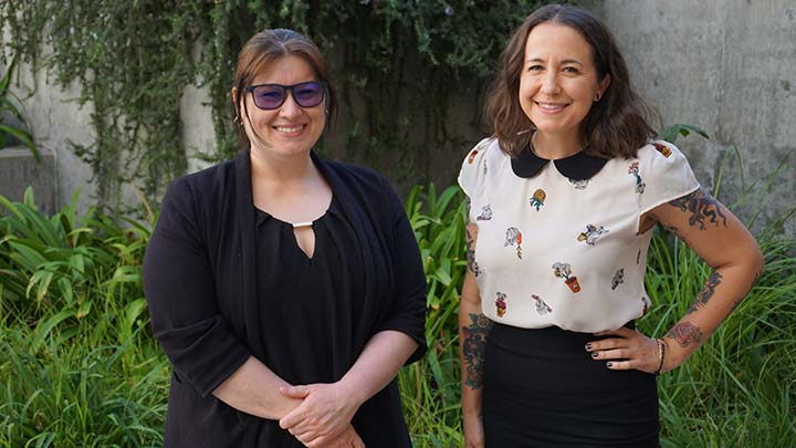 Two women smile and pose in front of a garden of grasses and plants.