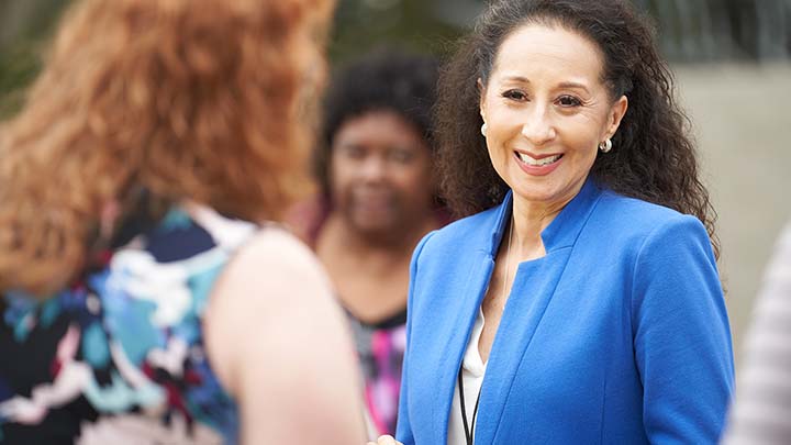 A Hispanic woman with long, wavy hair, wearing a blue blazer is smiling, her hands brought together, and facing a redhaired woman in a print dress.