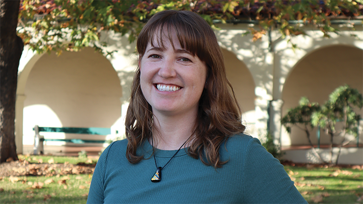 A young woman in a blue top is smiling broadly, her teeth showing. Her brown bangs hang down to her eyebrows and she is wearing a triangle-shaped, multicolored pendant. In the background is an arched building with a bench.