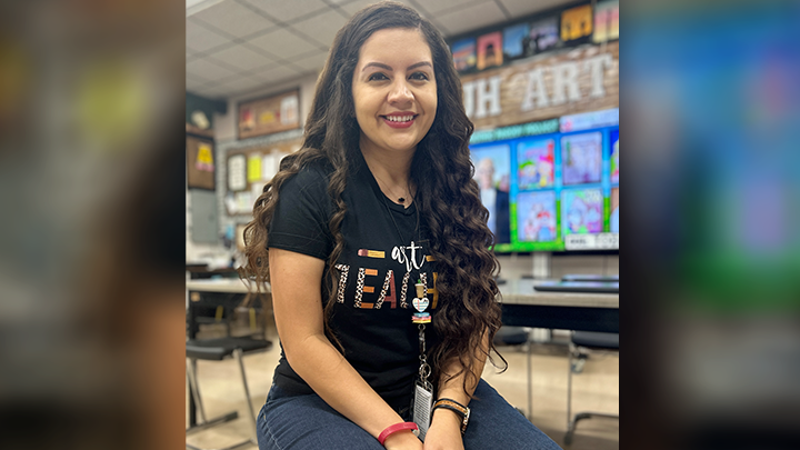 A smiling Amanda Casey is photographed sitting on desk in her classroom.