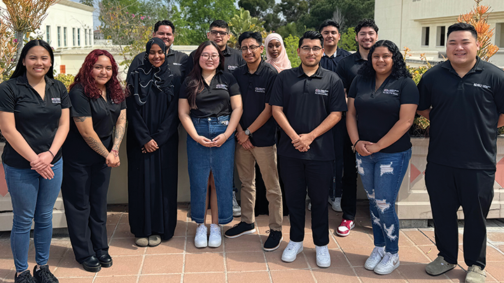 13 students smile for the camera over a picturesque background of the San Diego State University campus.