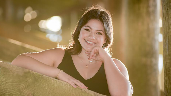 A sun-splashed photo of a smiling SDSU grad Aundrea Kaiser standing under a San Diego-area beach pier at sunset