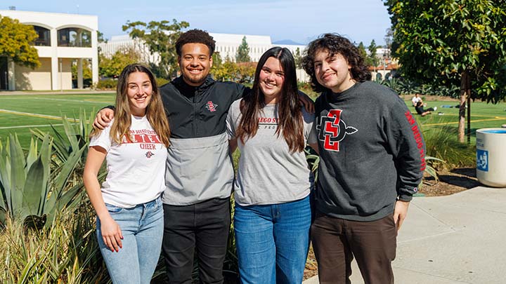 Four students, all wearing some type of SDSU gear, stand shoulder-to-shoulder on a campus path. Behind them is an open, green recreation field.