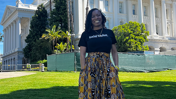 Maxine Amaru poses in outside California's capitol building wearing a long colorful skirt and a black shirt that reads "social worker"