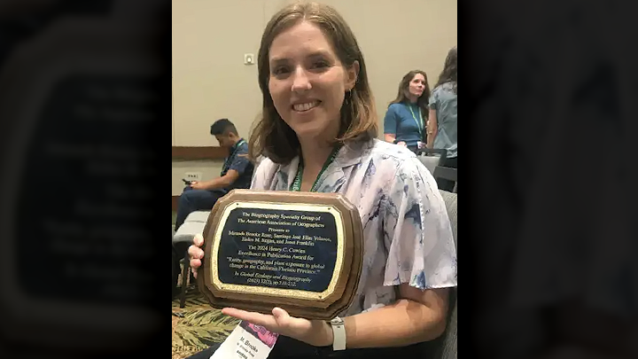 A woman with shoulder-length brunette hair, wearing a short-sleeved blouse, is smiling and seated in a meeting room and holding a plaque in front of her.