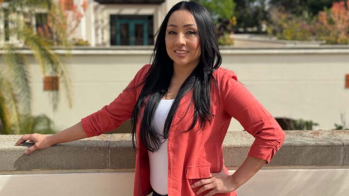 A young woman with long black hair, wearing a coral-colored jacket over a white top, is standing at a wall overlooking a courtyard. Her right arm is stretched along the top of the wall and her left hand is on her hip.