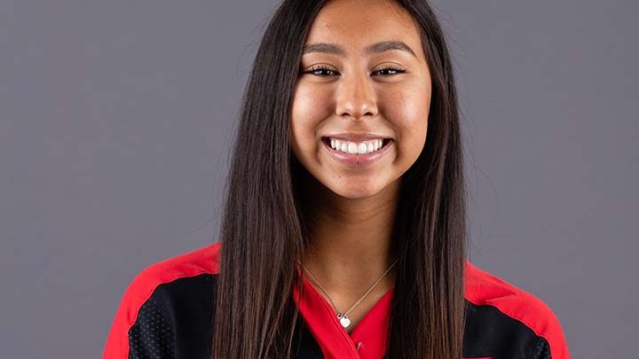 A portrait of a young woman with long, sark brown hair falling below her shoulders. She is smiling broadly, wearing a red-and-black top and a pendant around her neck