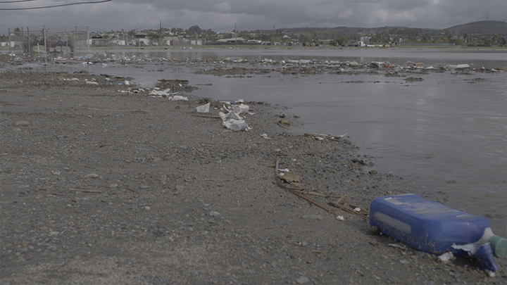 The gray, rocky shoreline of a shallow river, with trash and debris along the water's edge. On the opposite side of the river are hills and unidentifiable structures.