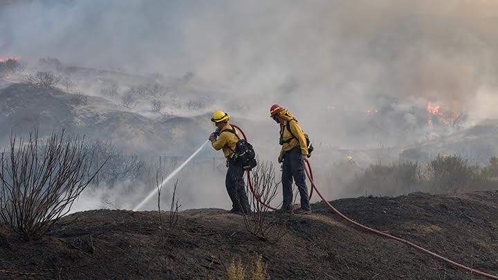 Two firefighters are seen on a smoky, burned landscape. One is direct a strong stream of water from a fire hose toward a section of ground obscured from view. There is smoke everywhere and some hills and flames in the background.