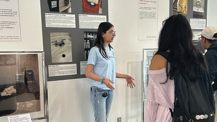 A young woman in a blue blouse and faded blue jeans, both arms extended in front of her, is standing in front of a display of photographs and text posted on a wall, speaking to two people with their backs to the camera. One photo shows a sink.