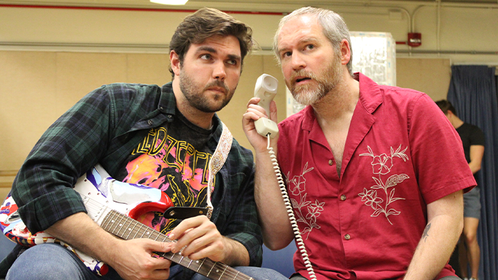 Two cast members pose for a photograph with a prop phone and guitar