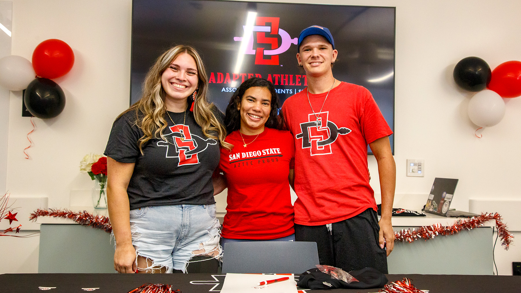 Three young women and a young man are standing side by side in an office with a sign behind them, partially obscured, that reads Adapted Athletics, with a San Diego State interlock logo above it.
