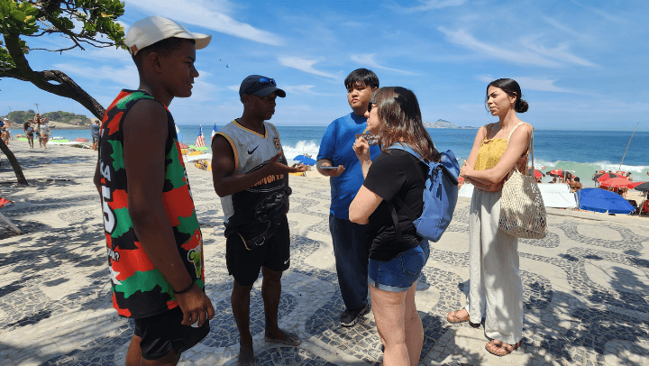 On a sandy beach with blue water in the background and a brilliant blue sky overhead, three students are speaking with two local residents 