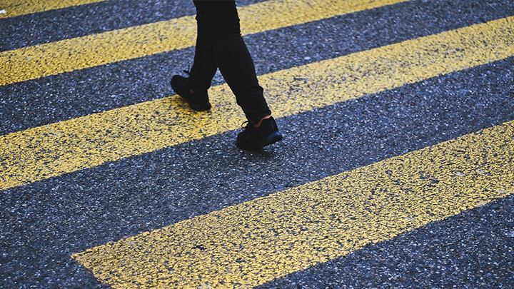 The two legs of a pedestrian, seen only from the knees down, are seen on an asphalt street with yellow crosswalk symbols.