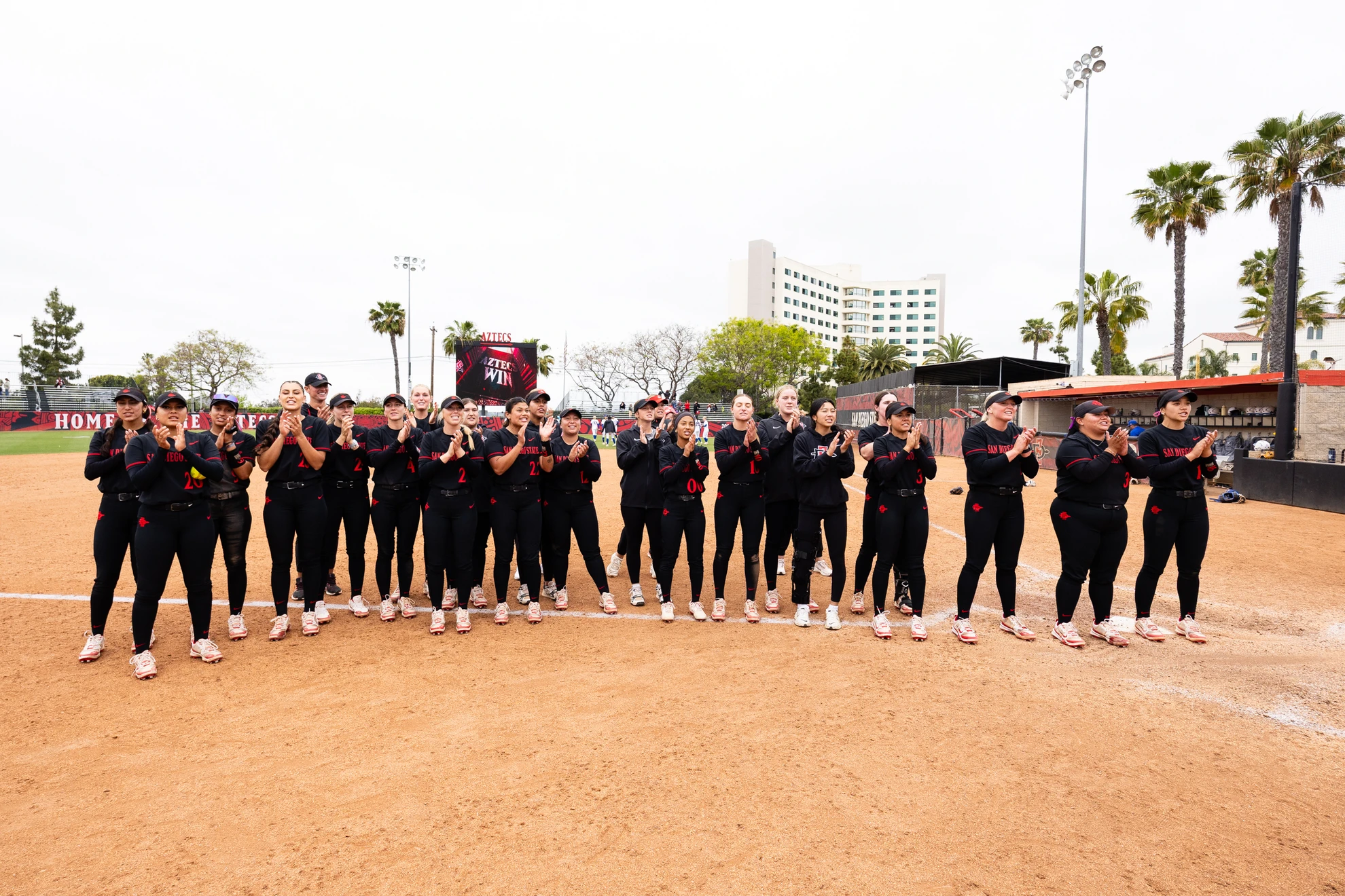 The SDSU softball team stand along the third base line applauding the fans in the seats.