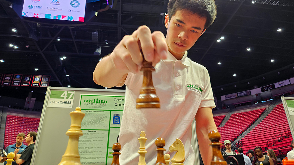 A young man in a white polo shirt with TEAM CHESS printed on the upper left holds a wooden chess piece as if about to position it with other pieces on a chessboard. The background depicts people milling about in a sports arena.