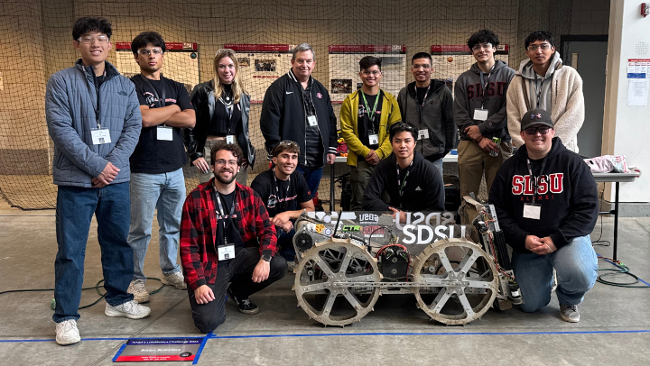Eleven students and a faculty member are lined up in rows of eight and four behind a metallic rover. Visible are two wheels with open spokes and a see-through panel with SDSU imprinted on it.
