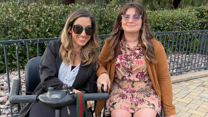 On the left, a woman seated in a motorized scooter wearing big sunglasses and a dark jacket over a blue striped top. On the right, a woman seated in a wheelchair, wearing a floral print dress. Behild them are a railing and some green shrubbery.