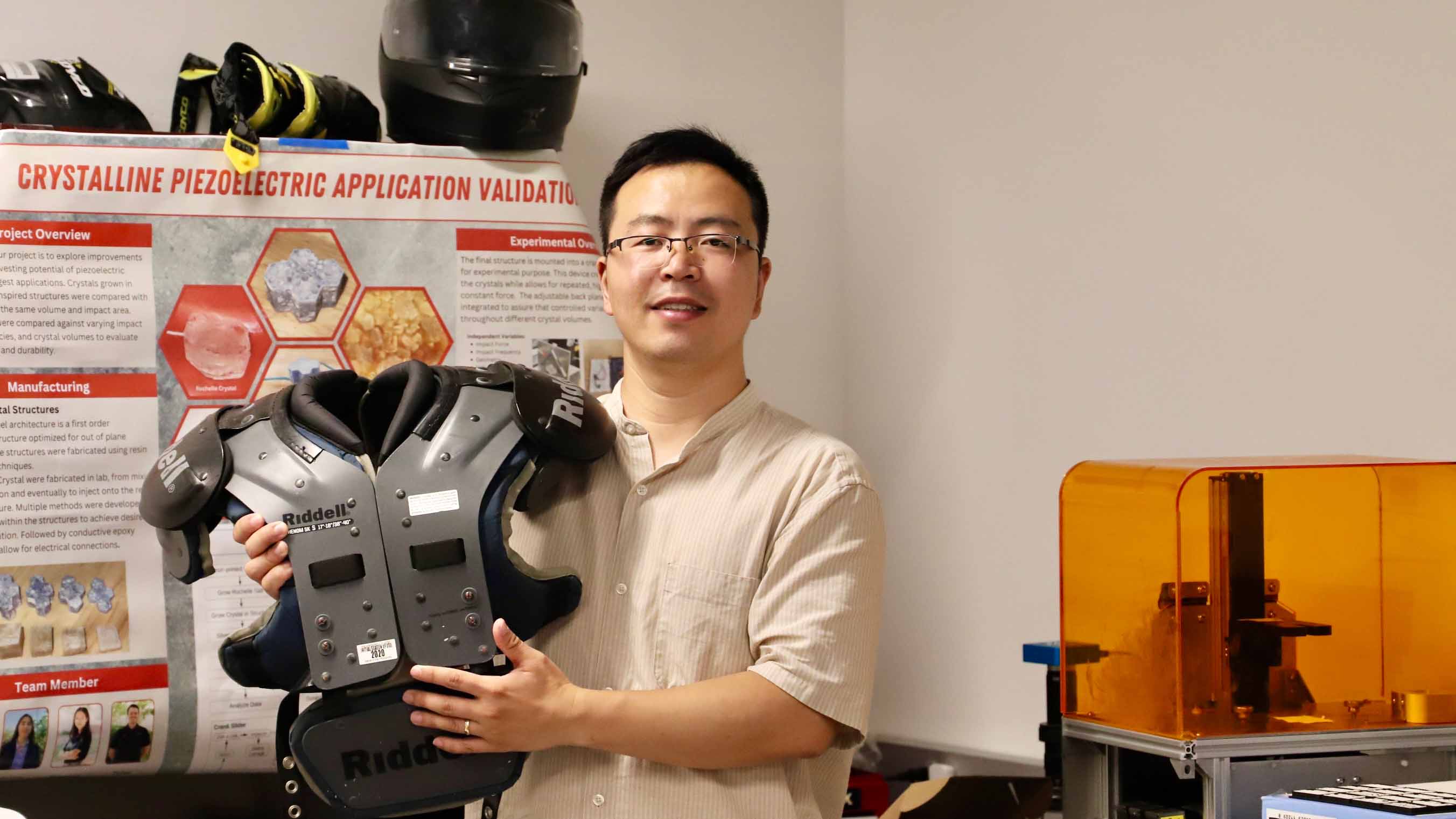 A man with short black hair and glasses wearing a beige, short-sleeve shirt holds an armored breastplate in a lab, with the poster for a science research project on the wall behind him.