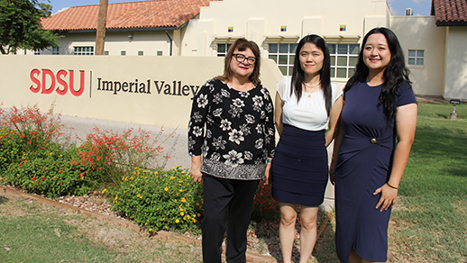 Three women stand in front of a monument sign reading SDSU Imperial Valley, with a one story building in the background and some flowers planted around the foot of the sign.