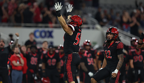 A football player in red and black jumps in celebration with his arms in the air as a player behind him looks on.