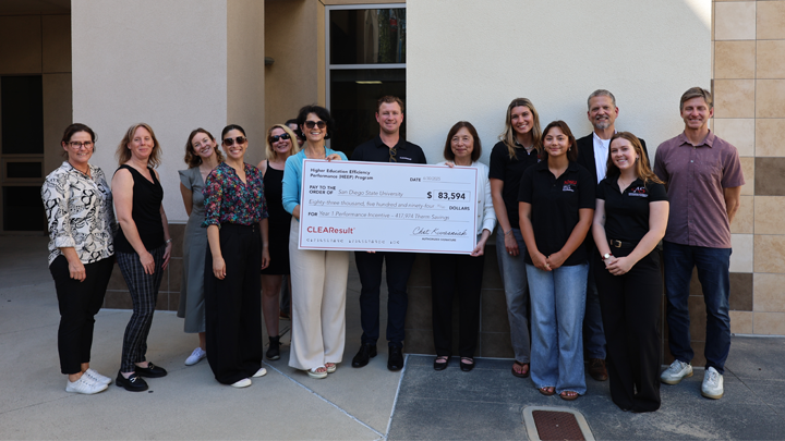 Thirteen men and women are lined up in front of a building with two women in the middle holding an oversized novelty check for $83,594, made out to San Diego State University. 