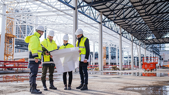 Three men and a woman at a construction site are examining a large sheet of white paper being held up by the woman. They are wearing reflective vests and hardhats and are standing amid open scaffolding, with a puddle of water to their left.