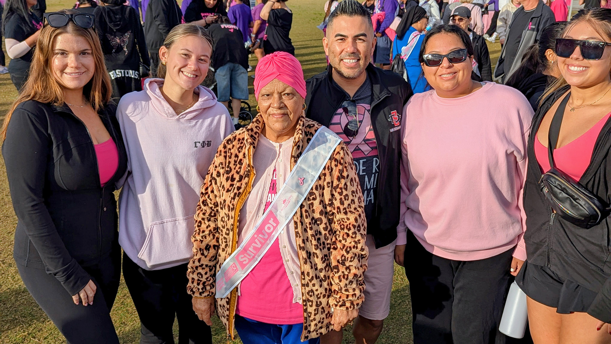 Six people stand side by side on a field with dozens more millng about in the background. A senior woman with a sash reading survivor and a middle-aged man are flanked by four young women, all wearing clothes with at least some pink.