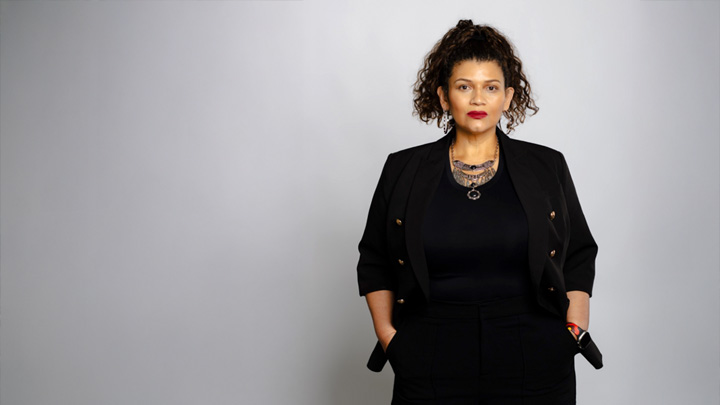 A woman dressed in black and wearing an elaborate silver necklace is standing in front of a stark gray wall with her hands in her pockets.