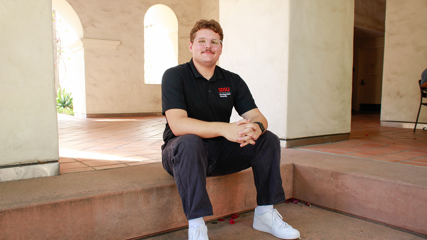 A young man sits on a short step outside a building, clasping his hands together between his knees. Behind him are arches in the building's architecture.