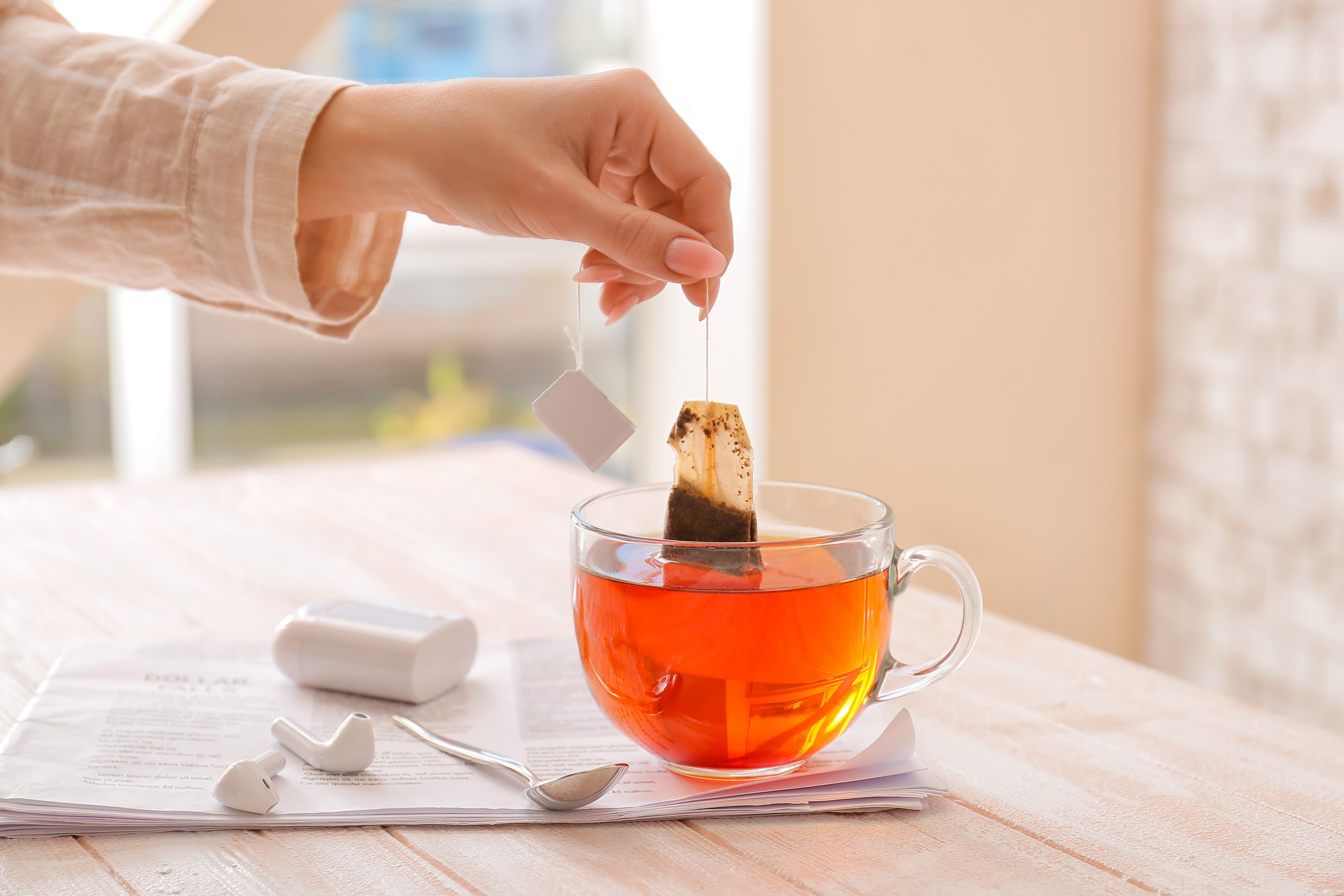 A closeup image of a woman's wrist and hand dipping a tea bag into a glass teacup with an orange beverage in it.