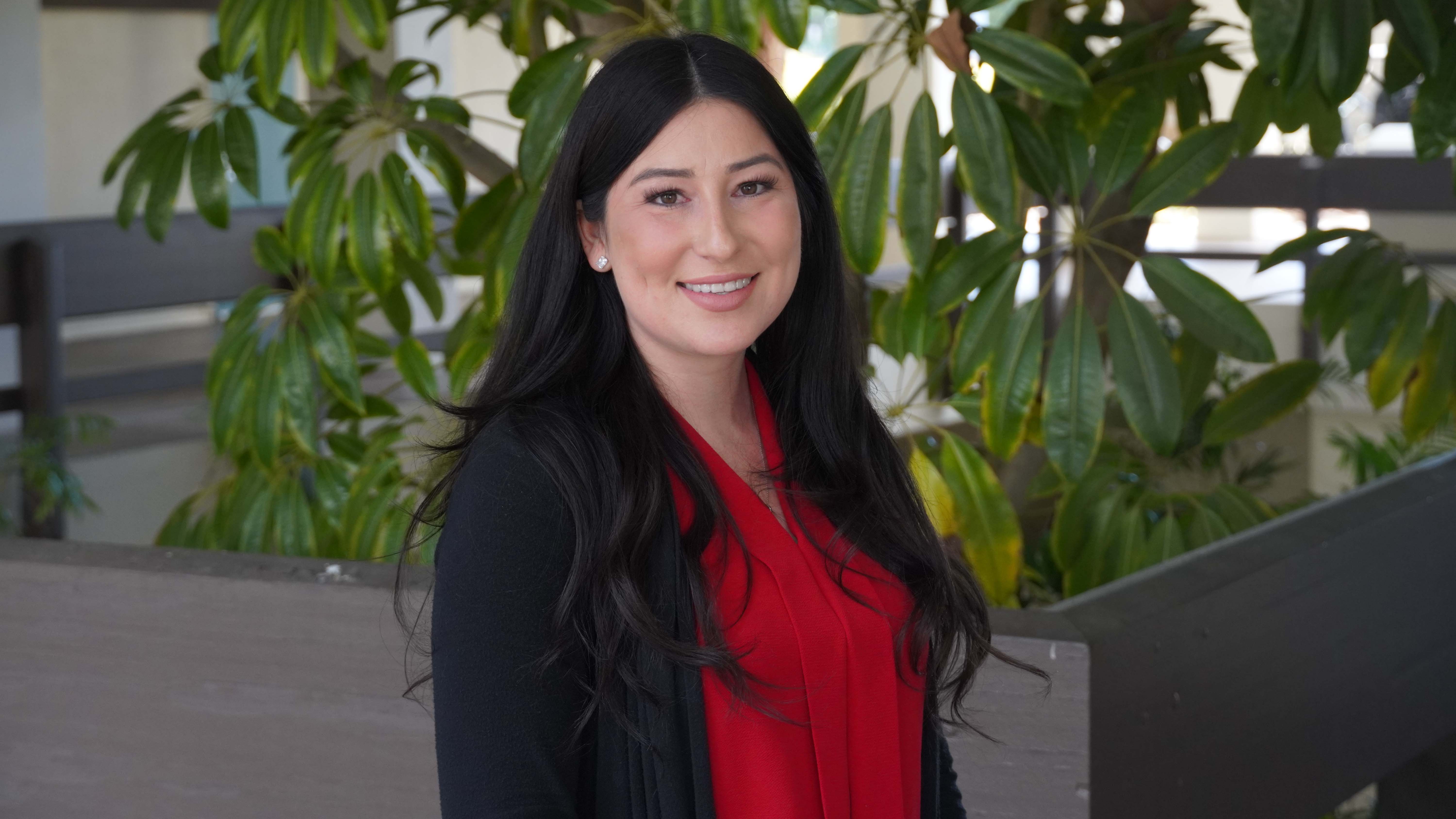 A woman dressed in a red top and black jacket faces the camera, smiling, with a green tree encaswed by a large planter behind her.