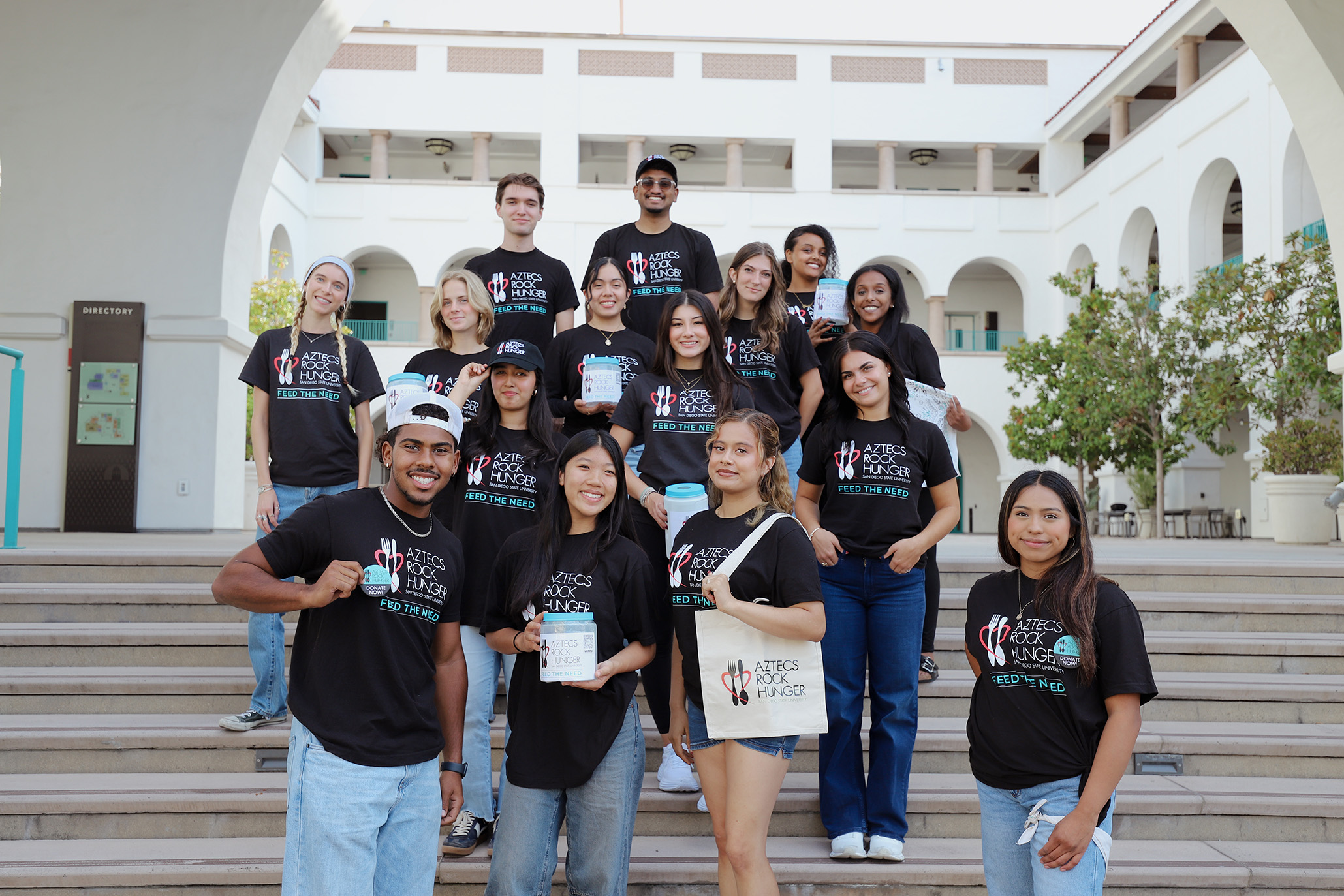 Fifteen young men and women, all wearing black t-shirts marked Aztecs Rock Hunger, stand in five rows of an outdoor staircase with a building in the background.