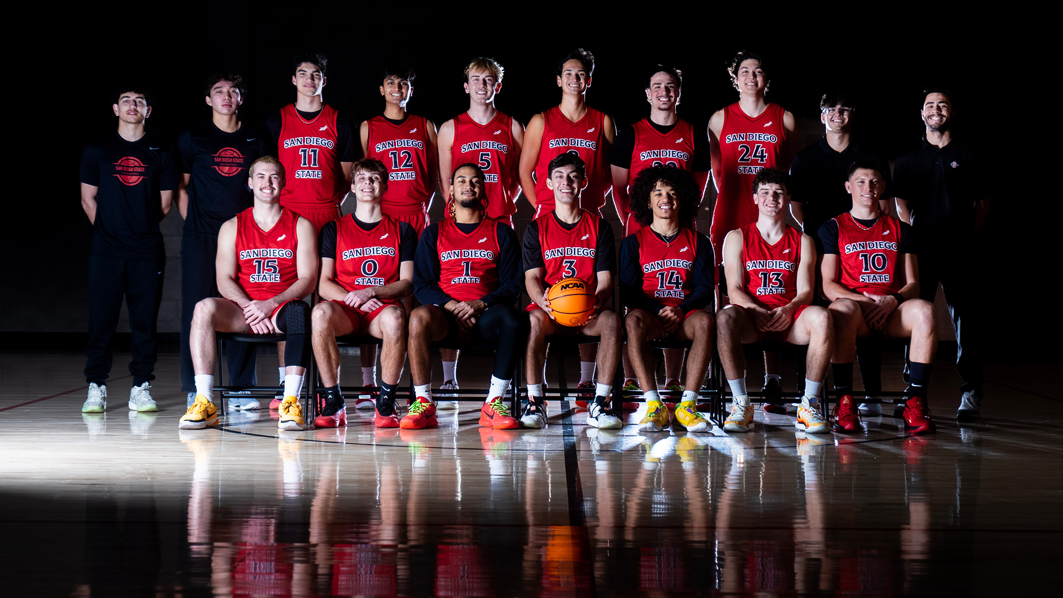 Two rows of young men in basketball uniforms, mostly with red jerseys. The bottom row is seated and reflected in the shiny surface of a black floor.