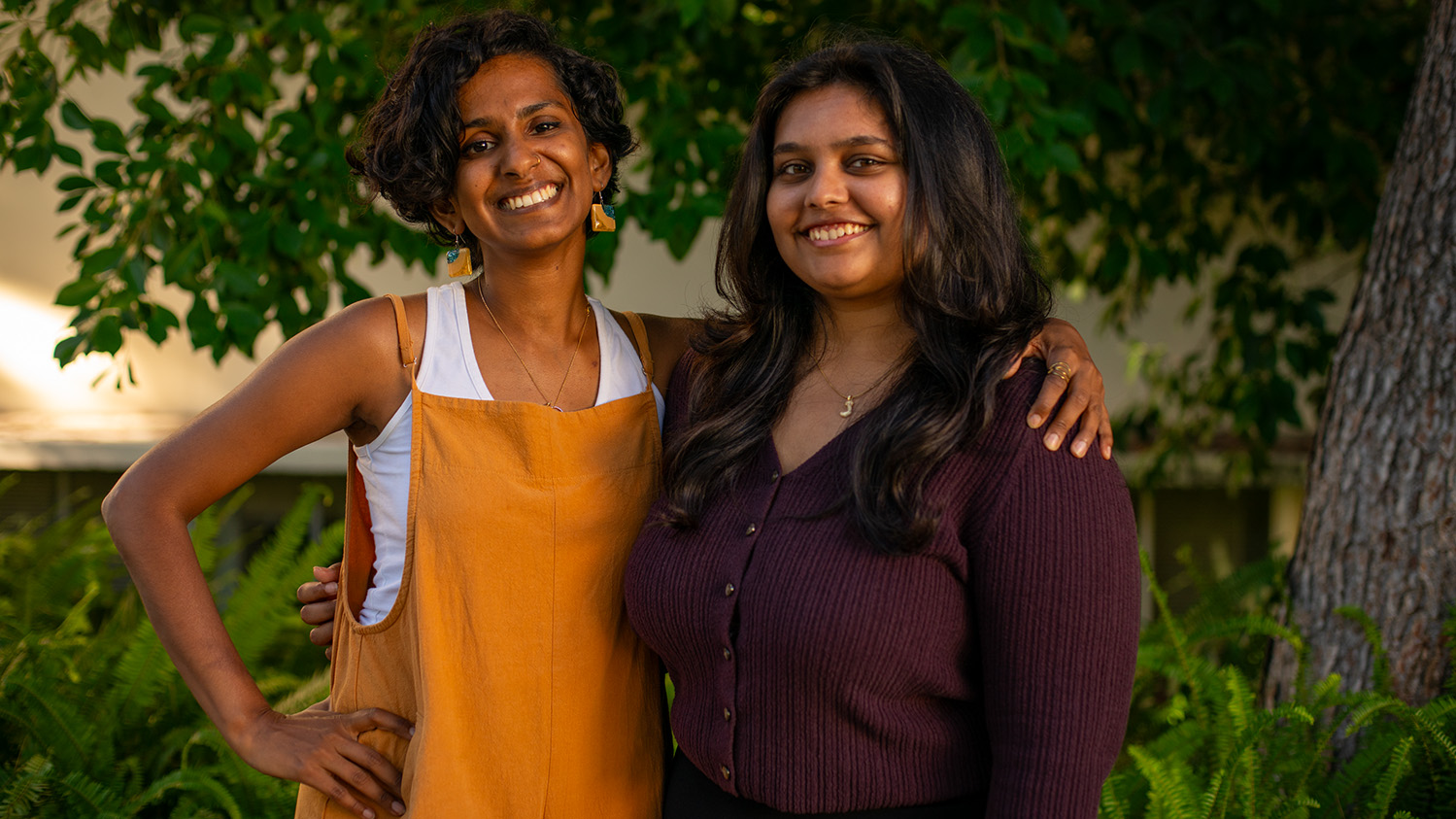 Two smiling women stand shoulder-to-shoulder. The one on the left has her arm around the other and they are stnding in front of a tree with lush green leaves and green landscaping.
