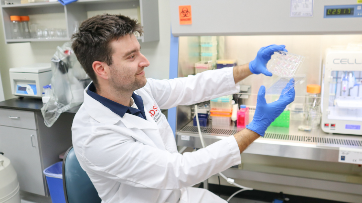 A young man seated in a laboratory and wearing a white lab coat and blue latex gloves holds a transparent lab instrument at arm's length.