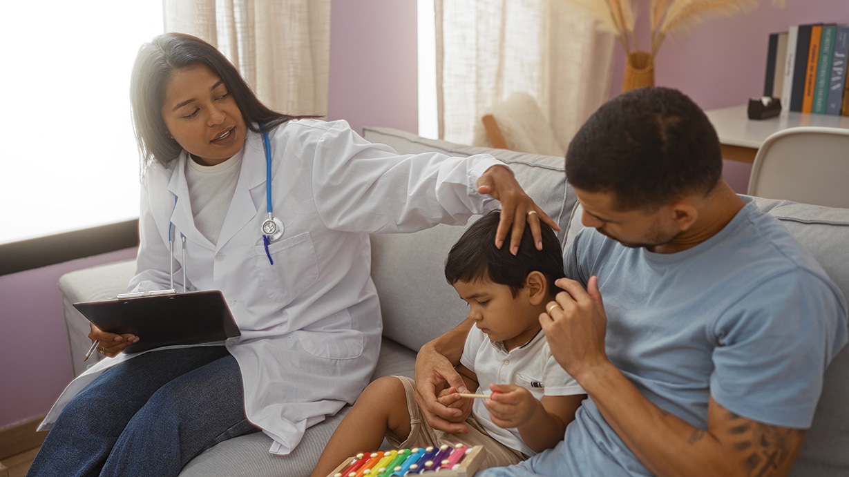 A young woman wearing a lab coat with a stethoscope around her nexh has her left hand on the head of a young boy playing with a toy xylophone as his father looks on.