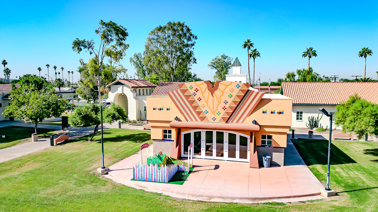 An aerial view of a campus with a one-story building in a Southwestern architectural style with large windows in front and patterns inspired by indigenous art. It faces a large green lawn.