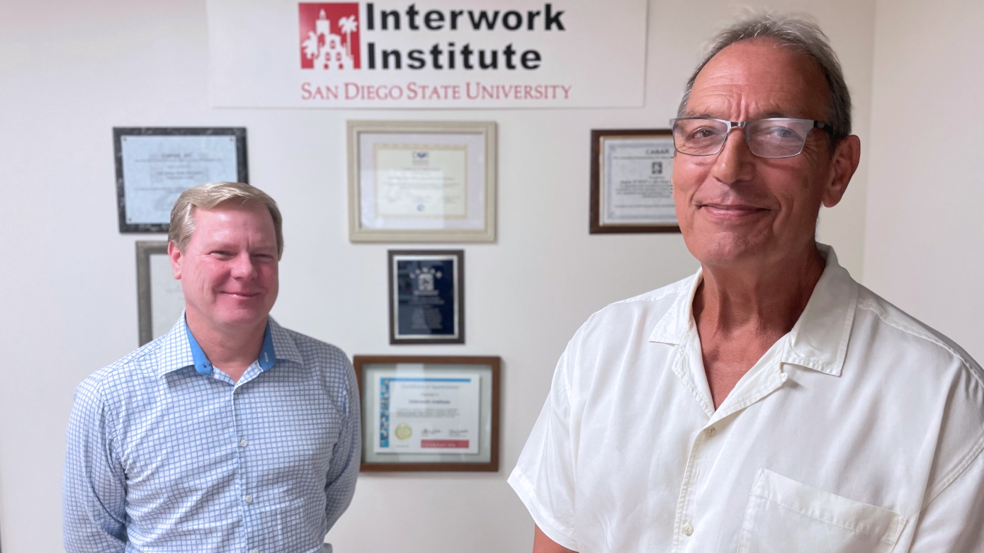 A man in a long sleeve blue checkered shirt and a man in a white short-slevve shirt are standing in front of a wall with six framed plaques or awards and a sign reading Interwork Institute San Diego State University.