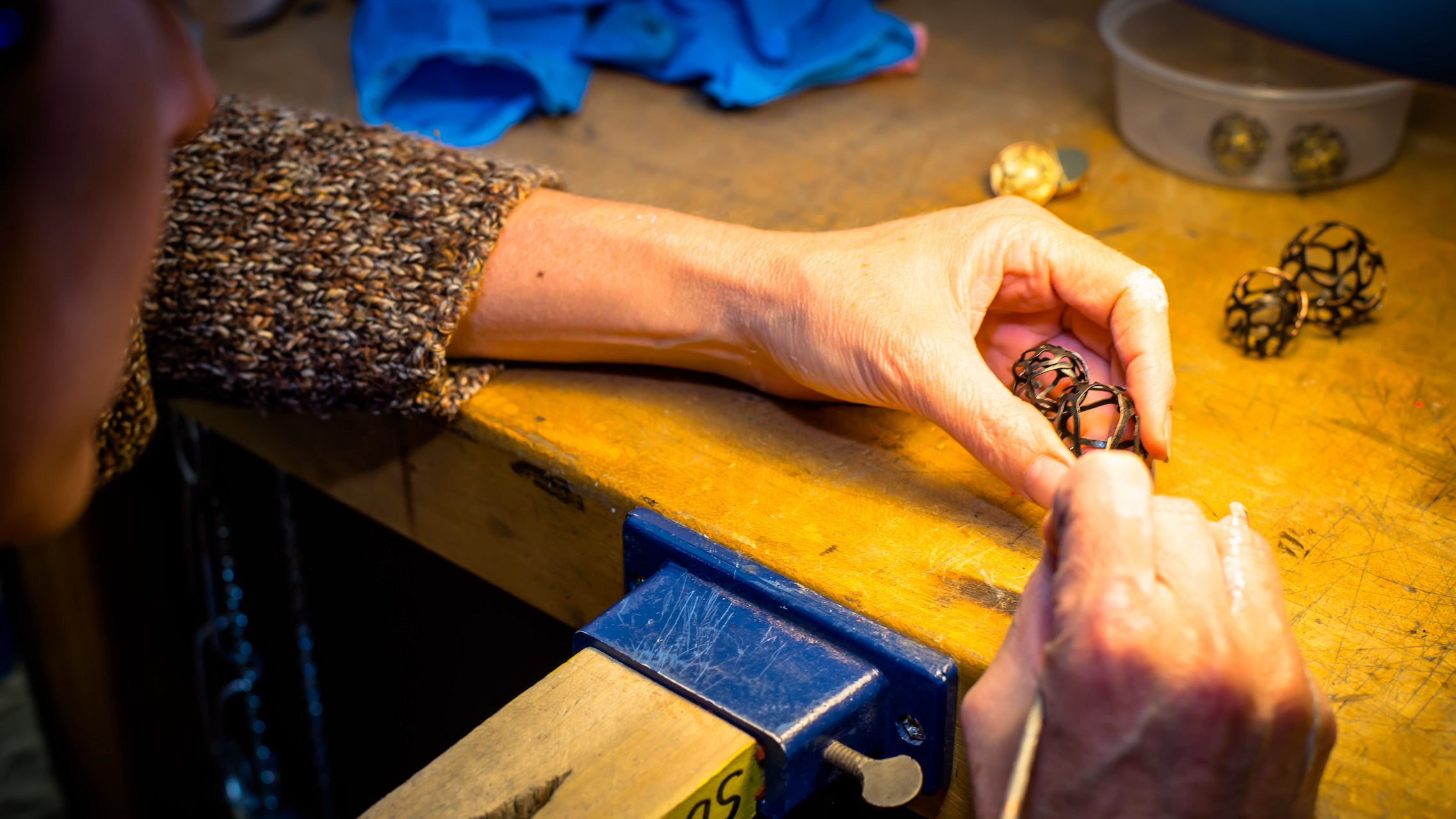 A closeup image of a pair of hands working with a pen-like device on a small piece of metalwork shaped like two open spheres.