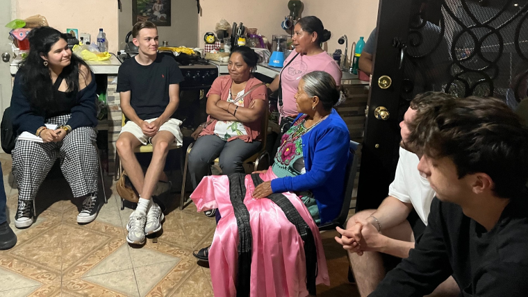 Inside a home, four students sit in a semicircle arounf three women, the oldest of which has a swath of pink fabric draped across her lap.
