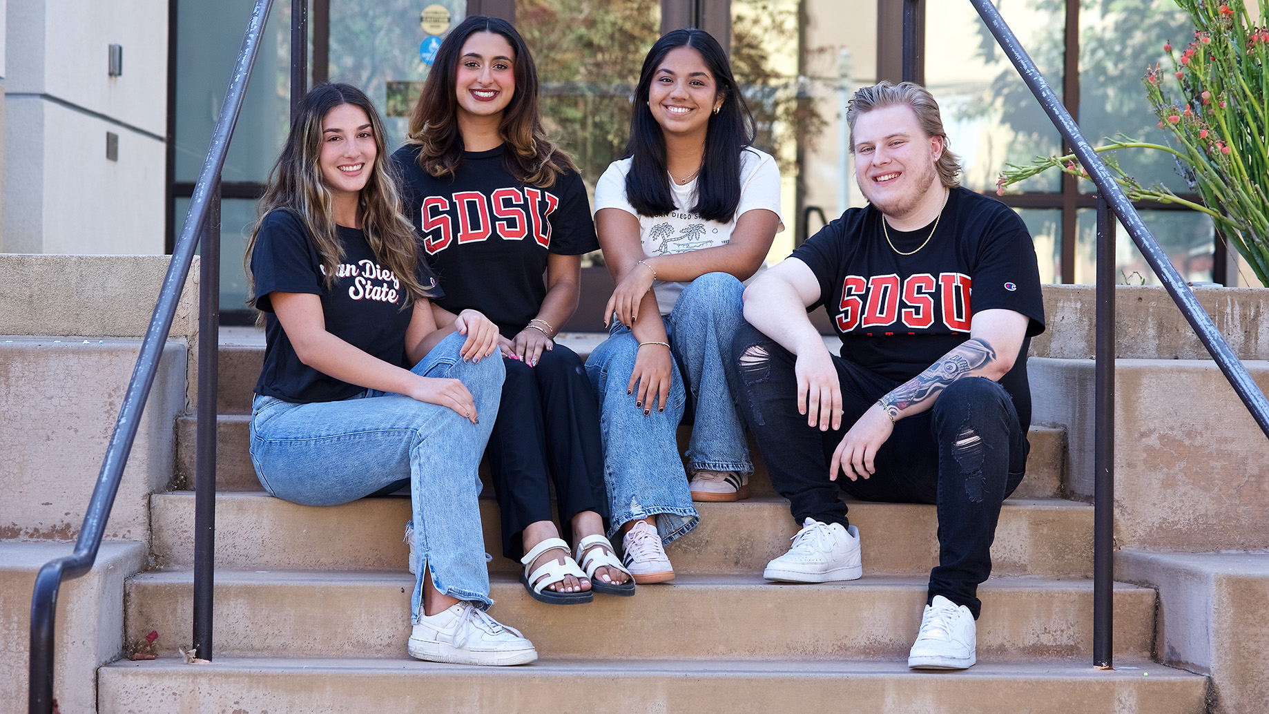 Four students, three female and one male, are sitting on a short, narrow staircase leading to a reflective glass door. All but one are wearing San Diego State University gear.