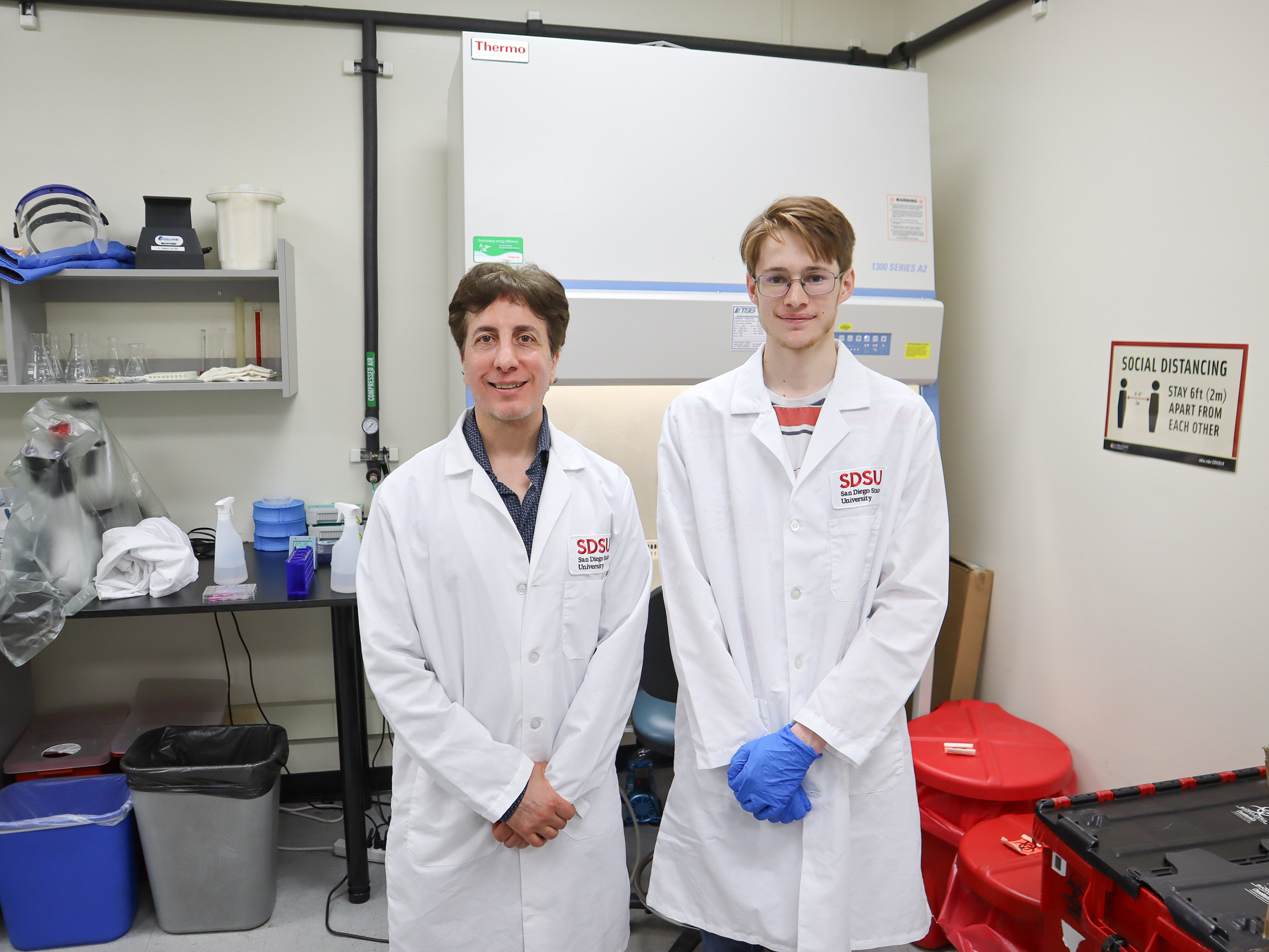 Two men wearing white lab coats pose in a laboratory, their hands clasped in front of them, with assorted material on a shelf behind them.